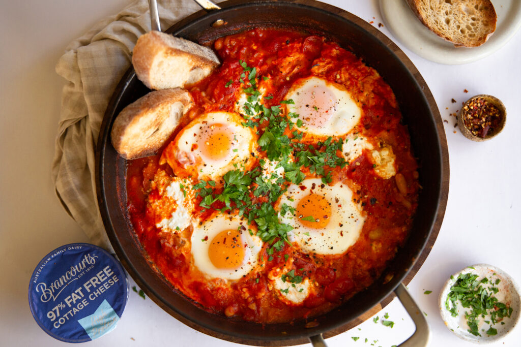 Smoky White Bean Shakshuka in a sizzling pan with rich tomato sauce, creamy cottage cheese, and perfectly poached eggs.
